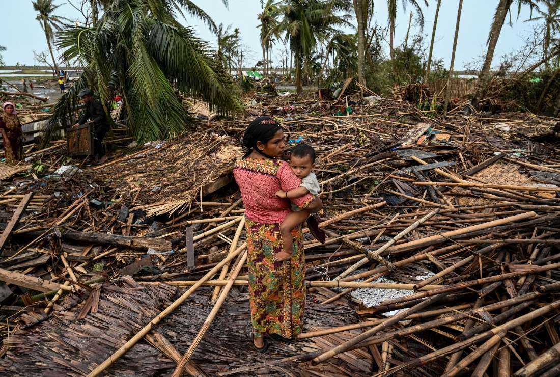 A Rohingya woman carries her baby next to her destroyed house at Basara refugee camp in Sittwe on May 16 after cyclone Mocha made landfall